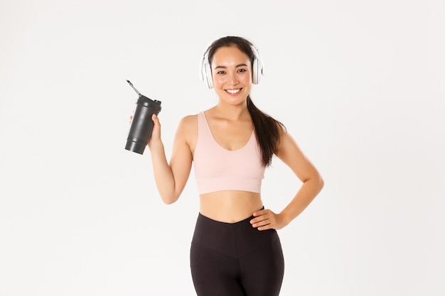 Sport, benessere e concetto di stile di vita attivo. Attraente ragazza asiatica fitness sottile e in forma in cuffia, ascolto musica durante l'allenamento, acqua potabile o proteine dalla bottiglia, muro bianco.