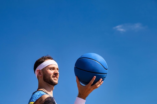 Sport e basket Giovane uomo che tiene palla da basket in braccio guardando lontano sorridente al cielo blu all'aperto