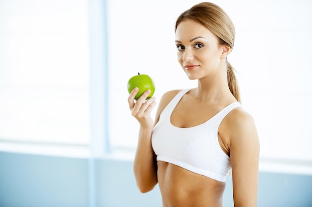 Sport e dieta. Bella giovane donna in abbigliamento sportivo che tiene mela verde e sorride alla macchina fotografica