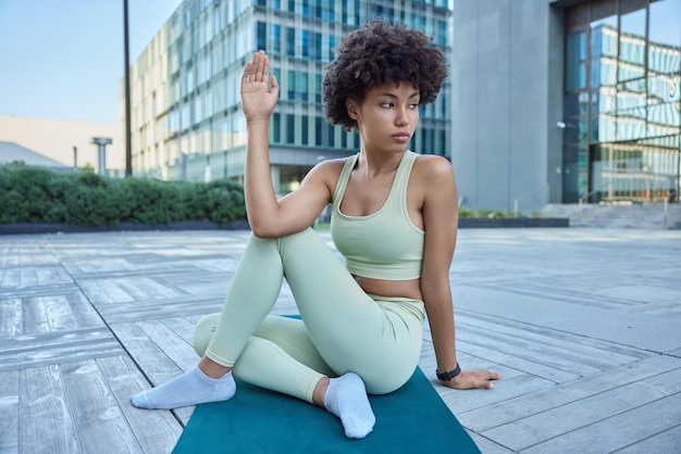 Sport fitness e concetto di allenamento. La giovane donna riccia premurosa allunga le braccia si allena sul tappetino vestito in pose di abbigliamento attivo all'aperto sullo sfondo urbano durante le pratiche diurne di stretching.