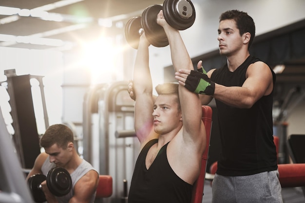 sport, fitness, stile di vita, powerlifting e concetto di persone - gruppo di uomini con manubri e personal trainer che flettono i muscoli in palestra