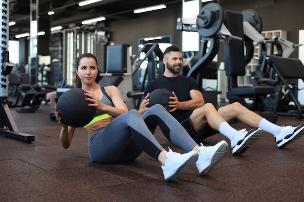 Sport giovani seduti sul pavimento di una palestra che si allenano insieme a una palla medica durante una sessione di esercizi addominali.