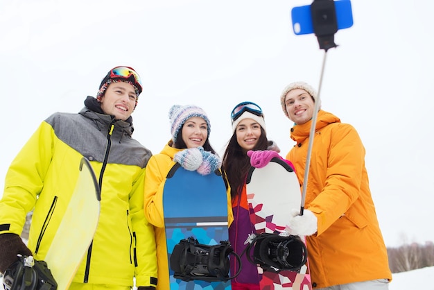 sport invernali, tempo libero, amicizia, tecnologia e concetto di persone - amici felici con snowboard e smartphone che prendono selfie