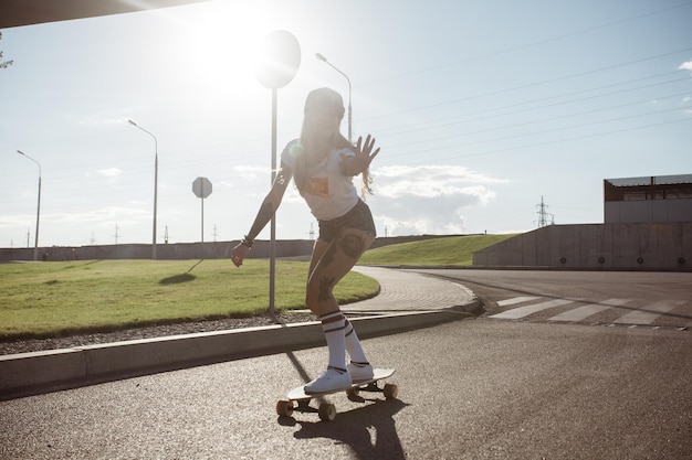 Sport, stile di vita, concetto estremo e persone - Tatuaggi di bella ragazza in sella a longboard sulla strada in città con tempo soleggiato. Ragazza hipster ritratto sorridente con un longboard al tramonto.