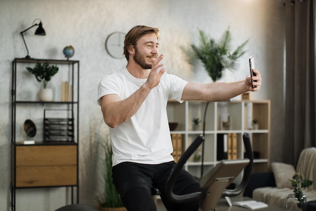 Sport uomo caucasico barbuto in abbigliamento sportivo con videochiamata online mentre si pedala in bicicletta