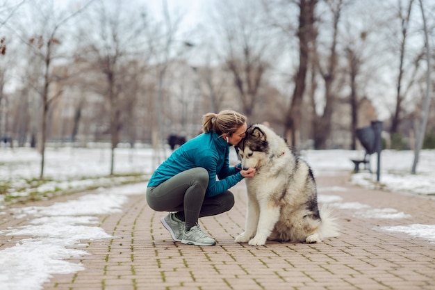 Sportiva che bacia il suo cane e lo abbraccia mentre è accovacciato nel parco con tempo nevoso. Animali domestici, attività ricreative, attività del fine settimana