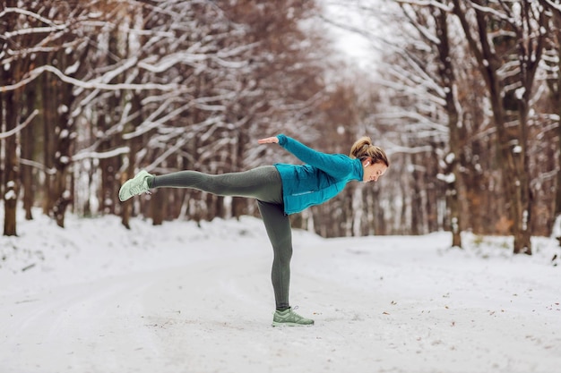 Sportiva in forma in equilibrio su un piede mentre si sta in piedi nella natura in una giornata invernale innevata. Fitness invernale, equilibrio, natura