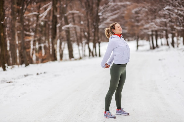 Sportiva in piedi in natura sulla neve in inverno e facendo esercizi di riscaldamento. Natura, foresta, fitness invernale, stretching