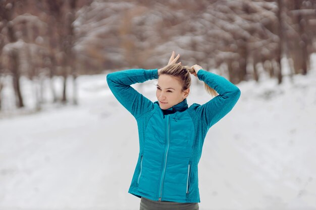 Sportiva in piedi nella natura con tempo nevoso e legare i capelli. Vita sana, sport invernali, acconciatura, neve