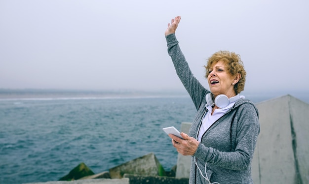 Sportiva senior che utilizza smartphone e saluta dal molo sul mare