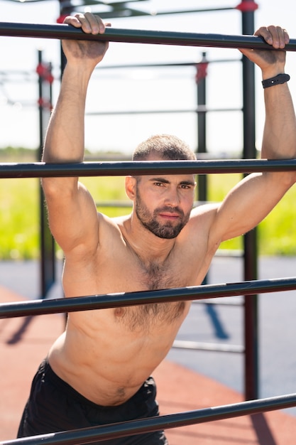 Sportivo a torso nudo forte di successo che ti guarda mentre ti appoggia alle barre dello sport durante l'allenamento all'aperto