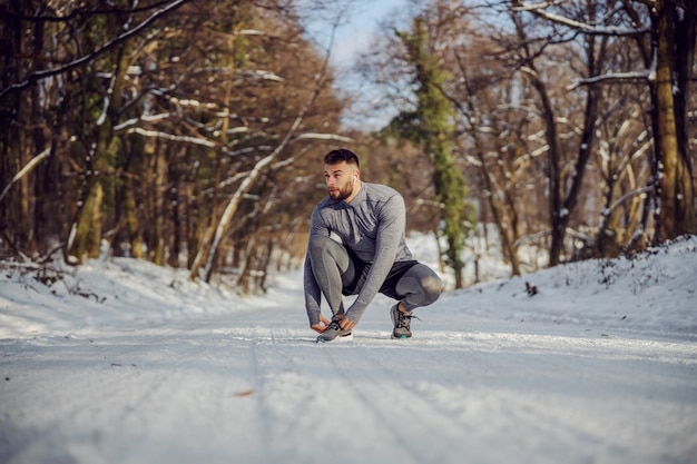 Sportivo accovacciato e legando i lacci delle scarpe in natura durante la soleggiata giornata invernale innevata. Abbigliamento sportivo, fitness invernale, clima freddo