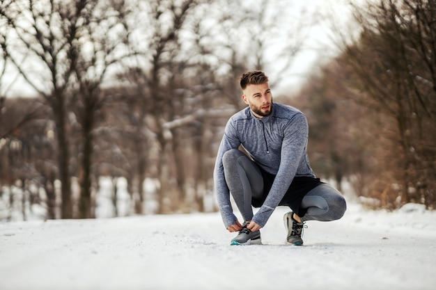 Sportivo accovacciato sul sentiero in natura e allacciare i lacci delle scarpe al giorno di inverno nevoso. Stile di vita sano, abbigliamento sportivo, tempo nevoso