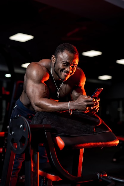 Sportivo africano con un corpo perfetto sorride e legge il messaggio in palestra. Sfondo chiaro scuro.