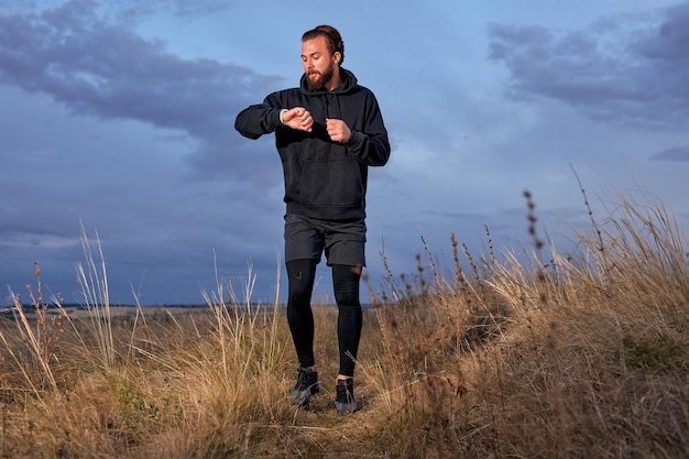 Sportivo che controlla l'ora su orologi da polso intelligenti mentre fa jogging all'alba al mattino all'aperto nella natura, sport all'aria aperta, allenamento. giovane ragazzo caucasico in abito sportivo nero