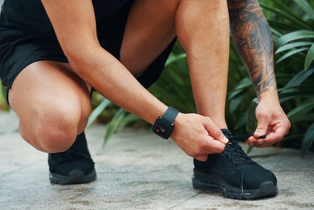 Sportivo con smartwatch che allaccia i lacci delle scarpe quando si prepara per fare jogging all'aperto