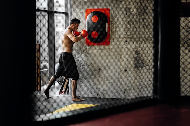 Sportivo con un torso nudo e guanti da boxe rossi colpisce l'attrezzatura per la boxe su un muro di cemento in palestra dietro la rete.