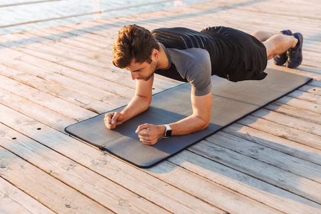 Sportivo concentrato in tuta da ginnastica che si trova sulla stuoia e che fa plancia mentre risolvendo sul pilastro di legno alla spiaggia nella mattina