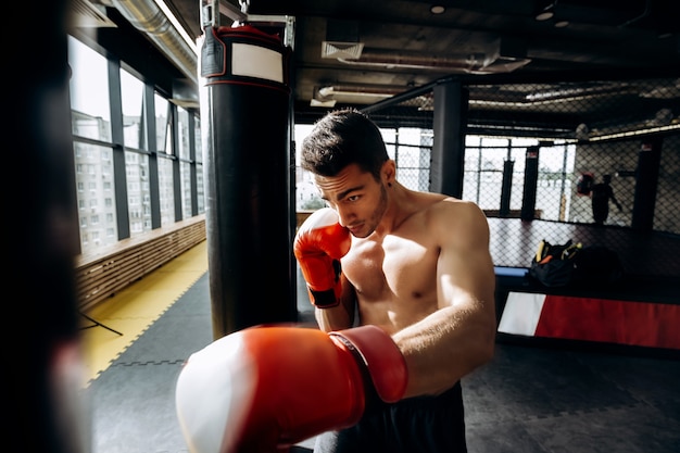 Sportivo in guanti da boxe rossi con un torso nudo colpisce il sacco da boxe in palestra sullo sfondo del ring.