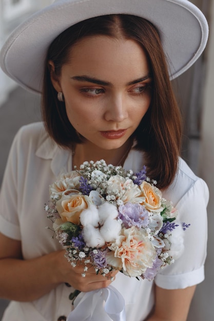 sposa alla moda con bouquet da sposa elegante di fiori e in cappello bianco che distoglie lo sguardo