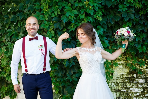 Sposa allegra delle coppie in un vestito bianco con un mazzo mentre lo sposo con le bretelle e la cravatta a farfalla che tengono le mani. Sullo sfondo di un muro con foglie verdi. Coppia felice. concetto di matrimonio.