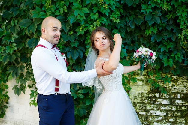 Sposa allegra delle coppie in un vestito bianco con un mazzo mentre lo sposo con le bretelle e la cravatta a farfalla che tengono le mani. Sullo sfondo di un muro con foglie verdi. Coppia felice. concetto di matrimonio.