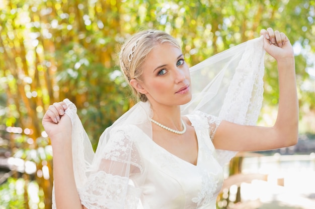 Sposa bionda contenta graziosa che tiene il suo velo fuori che sorride alla macchina fotografica