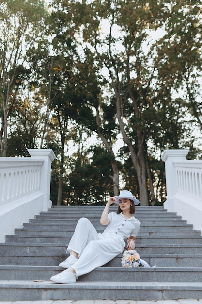 sposa con cappello bianco e costume seduta sulle scale con bouquet di fiori nel parco estivo