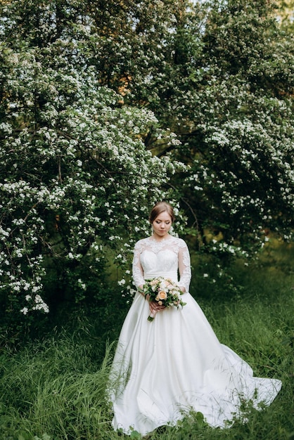 Sposa con un bouquet da sposa nella foresta vicino ai cespugli che sbocciano con fiori bianchi