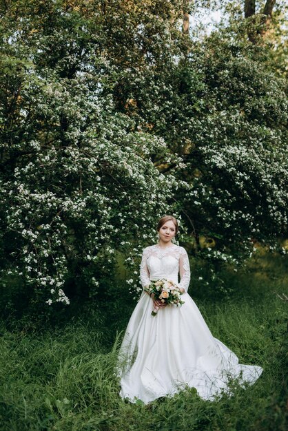 Sposa con un bouquet da sposa nella foresta vicino ai cespugli che sbocciano con fiori bianchi