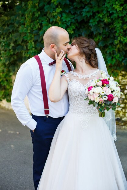 Sposa coppia allegra in un abito bianco con un bouquet mentre lo sposo con bretelle e papillon. Sullo sfondo di un muro con foglie verdi. Coppia felice. Il concetto di matrimonio.