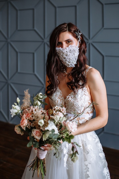 Sposa dentro con una maschera antivirale di nozze fatte a mano sul suo viso. Bouquet da sposa in mano