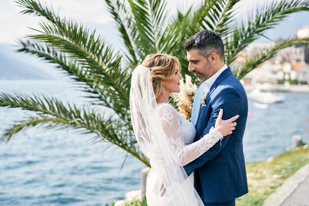 Sposa e sposo abbracciati che si guardano negli occhi vicino a una palma vicino al mare
