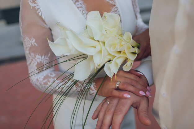 Sposa e sposo che si tengono per mano fedi nuziali e dettaglio del bouquet delle spose