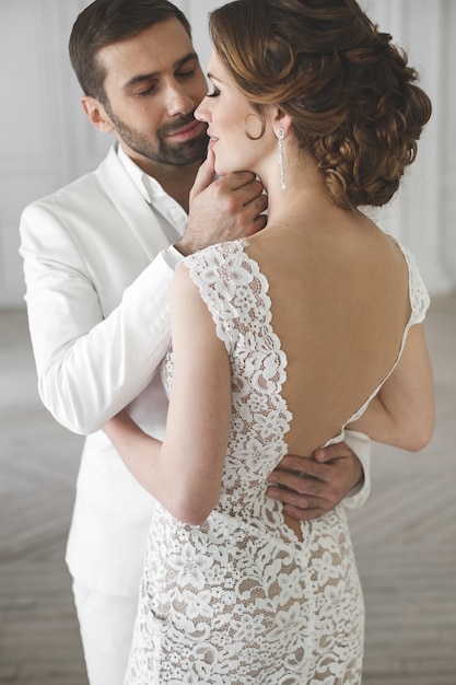 Sposa e sposo chic sposi in posa in uno studio bianco. Coppia felice ridendo. Vestito bianco. Abito bianco. Gioventù. Nozze. Lampadario. Divano. La porta.