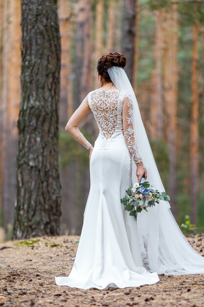 Sposa elegante in un abito da sposa con pizzo in piedi nei boschi tenendo bouquet da sposa di fiori