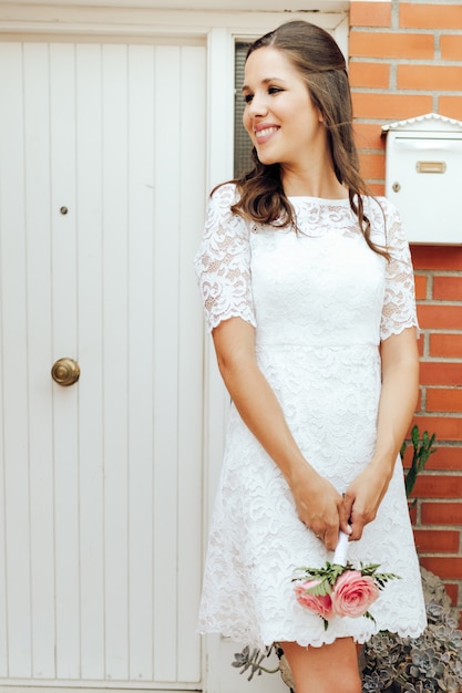 Sposa felice che tiene il suo piccolo mazzo di nozze delle rose rosa in porta bianca. Concetto di giorno del matrimonio.