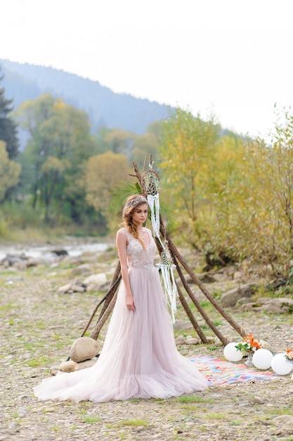 Sposa felice in un abito da sposa rosa. La ragazza tiene in mano un bouquet da sposa. Cerimonia di nozze in stile Boho vicino al fiume.