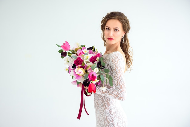 Sposa gioiosa della ragazza in un vestito lavorato a maglia bianco che posa con un mazzo di fiori