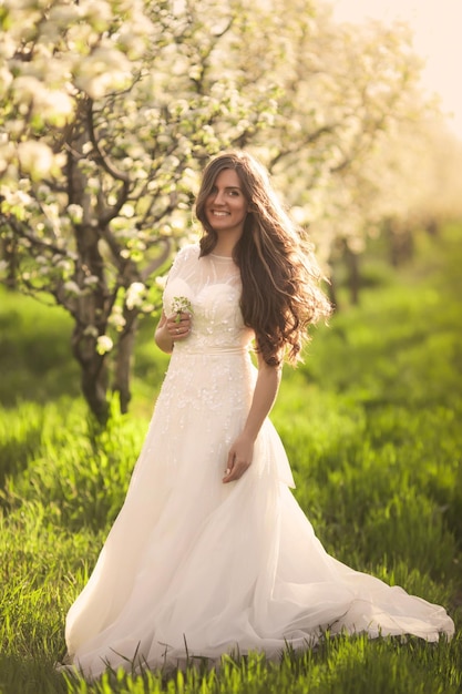 Sposa in abito da sposa avorio con lunghi capelli ricci che cammina nei giardini primaverili con alberi in fiore