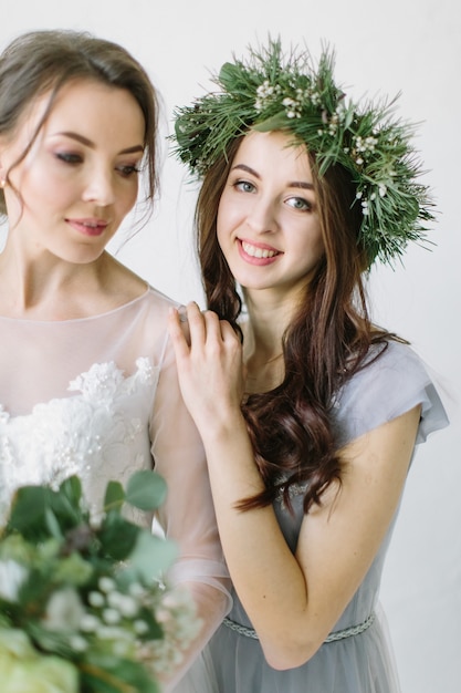 Sposa in abito da sposa bianco in piedi con la sua damigella d'onore sorridente in corona di pino e vestito blu