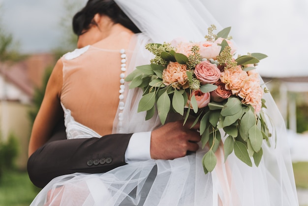 Sposa in vestito aperto indietro che abbraccia il suo sposo che sta tenendo un mazzo di nozze all&#39;aperto