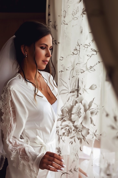 Sposa sveglia della giovane donna in accappatoio domestico alla finestra in casa di campagna. Mattina della sposa il giorno del matrimonio. La femmina felice sta aspettando di incontrare il suo sposo. Concetto di felice e lusso sposato. Copia spazio per il sito