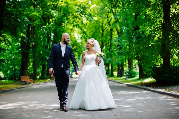 Sposi, bella giovane sposa e lo sposo in piedi in un parco all'aperto tenendosi per mano e sorridendo