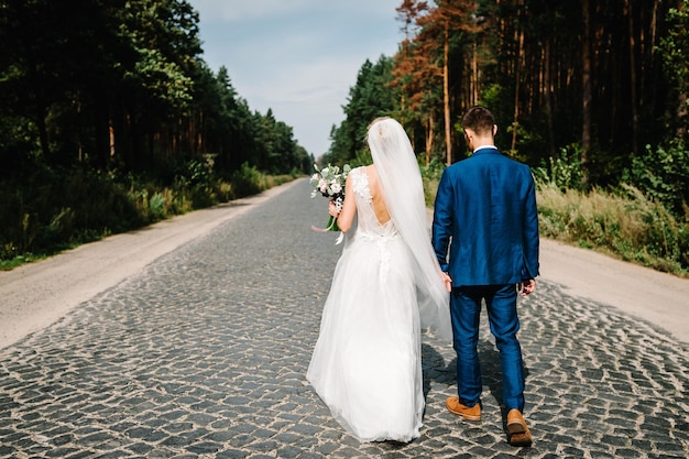 Sposi che camminano nella natura La sposa con il bouquet da sposa e lo sposo vanno sulla strada lastricata e si tengono per mano durante una passeggiata lungo la foresta Sposi della vita all'aria aperta Vista posteriore