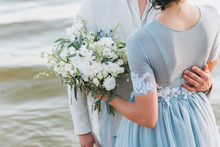 Sposo avendo la sua mano sulla vita della sua sposa, in piedi su una spiaggia. La sposa è in possesso di un bouquet.