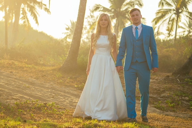 Sposo bello in un abito elegante e una sposa affascinante in un bellissimo abito da sposa sorridente sullo sfondo delle palme. concetto di una cerimonia di matrimonio chic e ricca sulla spiaggia