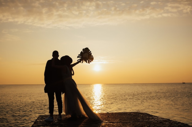sposo e sposa con bouquet in posa vicino al mare sul bellissimo tramonto
