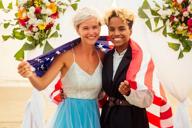 Sposo femminile afroamericano in abito nero e sposa bionda caucasica felice che ama insieme avvolta nella bandiera americana sulla cerimonia sulla spiaggia sotto l'arco floreale