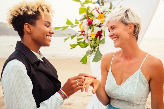 Sposo femminile afroamericano in abito nero e sposa bionda caucasica felice che ama insieme sulla cerimonia sulla spiaggia sotto l'arco floreale